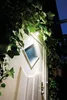 A white door with a diamond-shaped window, illuminated by a light above, surrounded by green ivy. Voir les images du produit
