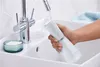A person is rinsing a white oral irrigator under a faucet in a bathroom sink. Podgląd zdjęcia produktu