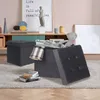 Two dark gray storage ottomans are shown on a gray rug in a room with a wooden floor. One ottoman is closed, and the other is open, revealing a teddy bear, a blanket, and some books. A shelf unit with decorative items and a framed picture is in the backgro Produktbild-Vorschau