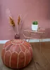 A Moroccan leather pouf is placed on a wooden floor next to a small side table with a cactus and a vase with dried flowers. Podgląd zdjęcia produktu