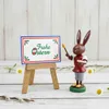 Bemalte Holzfigur Hase steht auf grünem Sockel trägt rotes Oberteil graue Hose gelbe Schuhe hält Pinsel und Palette neben Holzstaffelei mit Schild Frohe Ostern weißer Hintergrund mit Zweigen und kleinen Blumen Produktbild-Vorschau