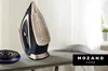 A cordless steam iron is placed on an ironing board next to a black accessory with blue light. Produktbild-Vorschau
