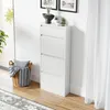 A white shoe cabinet with four flip-down doors stands against a white wall, next to a window with sheer curtains. A woven basket with a pillow and blanket sits on the floor to the left of the cabinet. Decorative plants and framed pictures are on top of the Náhľad obrázku produktu