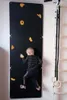 A young child is climbing on a black indoor climbing wall mounted on a white wall. A wooden ladder and gymnastic rings are visible to the right. Anteprima dell’immagine prodotto