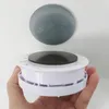 A hand holds a white circular device with a black disc on top, with a gray circular object floating above it against a white background. Produktbild-Vorschau