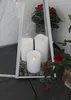 Three LED candles are placed inside a white lantern with gravel at the bottom. Red flowers and greenery are visible around the lantern. Produktbild-Vorschau