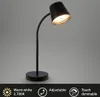 A black LED desk lamp with a flexible neck and a round base, shown against a plain gray background. The lamp is illuminated, casting a warm light. Náhľad obrázku produktu