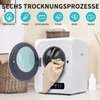 A white mini clothes dryer with its door open, showing a beige towel inside. The dryer is placed in a room with a basket of towels and a bottle of detergent nearby. Above the dryer, there are icons and text indicating different drying programs. Produktbild-Vorschau