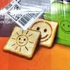 Two slices of toast with smiley faces are shown, one with a sun face and the other with a standard smiley face. The background is a gradient of brown, orange, and green. There is also a label with a barcode and text. Produktbild-Vorschau