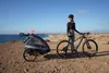 A woman with a bicycle and a child trailer on a beach with the ocean in the background. Produktbild-Vorschau