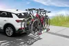 A white car with a bicycle rack attached to the rear, loaded with three bicycles, parked on a paved surface near a grassy dune and the ocean under a blue sky. Produktbild-Vorschau