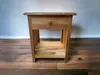 A wooden nightstand with a drawer and an open shelf, placed on a wooden floor against a light gray wall. Náhľad obrázku produktu