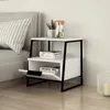 A modern white nightstand with black metal legs is placed next to a bed. The nightstand has two shelves, one with books and the other with a drawer that is partially open. A glass pitcher with water and lime, and a small potted plant are on top of the nigh Produktbild-Vorschau
