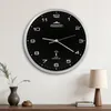A black analog wall clock with silver trim is mounted on a white wall above a wooden shelf. Anteprima dell’immagine prodotto