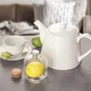 A white teapot, cup, saucer, spoon, macarons, lime, and a glass jar with a lemon are arranged on a gray surface, with a blurred living room in the background. Podgląd zdjęcia produktu