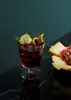 A glass of red vermouth with a cucumber slice and lemon peel garnish, next to a cheese and charcuterie board. Produktbild-Vorschau