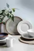 A collection of ceramic tableware, including plates, bowls, and a mug, arranged on a light-colored surface with a vase of eucalyptus branches in the background. Podgląd zdjęcia produktu