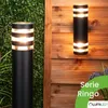 Two outdoor wall lights are shown, one mounted on a wooden fence and the other on a green hedge. The lights are cylindrical with horizontal bands of light. Anteprima dell’immagine prodotto
