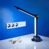 A desk lamp is positioned on a white surface in front of a blue wall with electrical outlets. Several small white components and a screwdriver are also on the surface. Produktbild-Vorschau