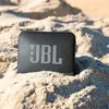 A black JBL speaker sits on a sandy beach. Voir les images du produit