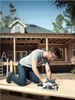 A man is kneeling on a wooden surface, using a cordless circular saw to cut a piece of wood. A house under construction is visible in the background. Anteprima dell’immagine prodotto