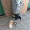 A stainless steel water heater with a shower fixture attached, placed on a speckled floor next to a wooden board and some firewood. There are also some plumbing fittings. Produktbild-Vorschau