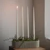 Four tall, thin white candles are arranged in a ribbed, rectangular holder filled with moss and small branches. One candle is lit. Produktbild-Vorschau
