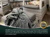 A grey blanket is draped over an ottoman in front of a wicker chair where a cat is sleeping. Produktbild-Vorschau
