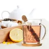 A glass tea infuser with a cork stopper, filled with tea leaves, sits next to a slice of lemon and a wooden tray filled with various tea bags and loose leaf teas. A white teapot is visible in the background. Produktbild-Vorschau
