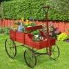 A red wooden wagon filled with gardening tools and plants sits on a green lawn in front of a hedge and a wooden fence. Podgląd zdjęcia produktu