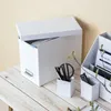 A white filing box and two smaller white containers are on a light wooden table. The filing box is open, showing files inside. One container holds scissors and pens, and the other holds pens. Small green branches are visible in the foreground and on the ri Produktbild-Vorschau
