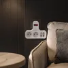 A white power strip with multiple outlets and an on/off switch is plugged into a dark wall, next to a chair and a round wooden table. Produktbild-Vorschau