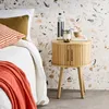 A bedside table with a candle and bottle of water next to a bed with pillows and a throw blanket. Produktbild-Vorschau