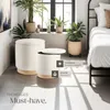 Two round storage ottomans are placed in a bright, modern room with plants and a bed in the background. Náhled obrázku produktu
