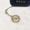 A close-up shot of a gold Guess logo charm with rhinestones, attached to a black strap, with the Guess logo visible on a black case in the background. Podgląd zdjęcia produktu