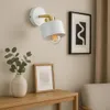 A modern wall lamp with a white shade and gold accents is mounted on a light beige wall above a wooden console table. A vase with eucalyptus branches and a framed picture are on the table. Náhled obrázku produktu