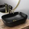 A black oval countertop sink with a gold faucet and drain, placed on a wooden surface with a gold-rimmed mirror in the background. Náhľad obrázku produktu