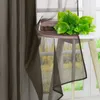 A sheer brown curtain is draped over a small round table next to a window. A bouquet of green artificial leaves is placed on the table. Produktbild-Vorschau