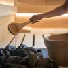 A person is pouring water from a wooden ladle onto hot stones in a sauna. Produktbild-Vorschau