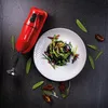 A red Bamix immersion blender is placed next to a plate of black pasta with green vegetables and sun-dried tomatoes, surrounded by snap peas and sage leaves on a dark textured surface. Podgląd zdjęcia produktu