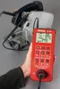 A hand holds a red electrical testing device next to a chop saw. Náhľad obrázku produktu