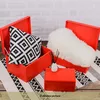 Three red storage boxes are arranged on a rug with a black and white geometric pattern. One box contains a black and white patterned pillow, another contains a fluffy white item, and a smaller box in front has a small white vase with reeds. Produktbild-Vorschau