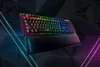 A gaming keyboard with RGB lighting and a wrist rest, set against a dark, geometric background with neon light accents. Produktbild-Vorschau