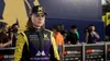 A female racing driver in a yellow and purple racing suit and cap stands on a podium with other people and cameras in the background. Produktbild-Vorschau