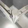 Close-up view of a corner joint of a white shower enclosure, showing screws and the white plastic frame against a gray concrete wall. Produktbild-Vorschau