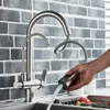 A hand is holding a kitchen faucet spray head, with water spraying out into a sink. The faucet is made of brushed metal and is mounted on a backsplash of gray brick tiles. Náhľad obrázku produktu