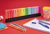 A collection of STABILO BOSS ORIGINAL highlighters in a desk set, along with colored pencils, a notebook, and a coffee cup, all arranged on a red surface. Anteprima dell’immagine prodotto