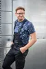 A man wearing black overalls and a blue camouflage t-shirt stands in front of a metallic wall. Náhled obrázku produktu