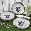 Several plates with a bull design are arranged on a grassy surface in front of a white wooden wall. A vase with greenery is also visible. Voir les images du produit