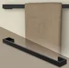 A black metal towel rack is shown mounted on a wall, with a beige towel draped over it. A reflection of the towel rack is visible on a glossy surface below. Produktbild-Vorschau